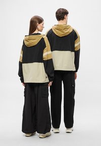 Μπουφάν με κουκούλα σε μαύρο, μπεζ και χρυσό, με χρωματικό μπλοκ και οριζόντιες ρίγες. Το μοντέλο φοράει φαρδιά μαύρα παντελόνια cargo και παπούτσια.