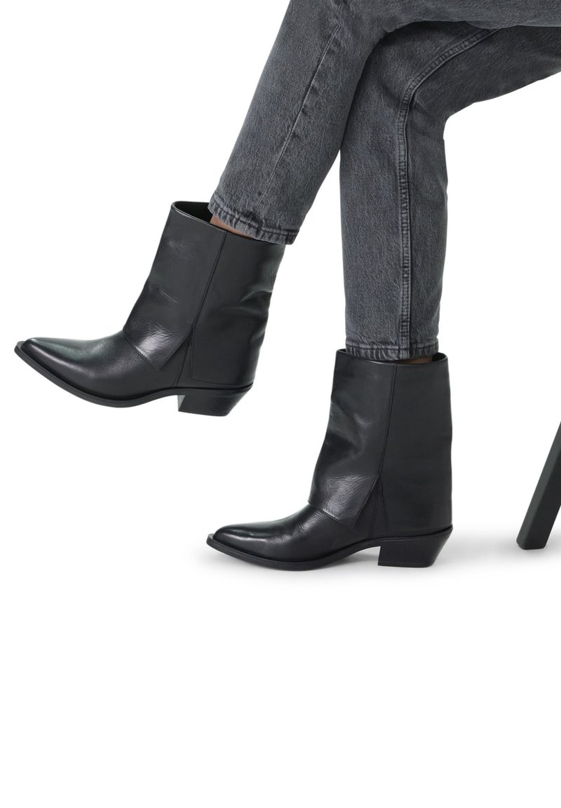 Stivaletti neri in pelle con punta affusolata, tacco basso a blocco e finitura liscia, mostrati con jeans grigi su uno sfondo bianco.