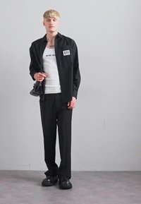 Personne blonde portant une chemise noire ouverte sur un débardeur blanc, un pantalon noir et des chaussures noires, tenant un petit sac noir d'une main.