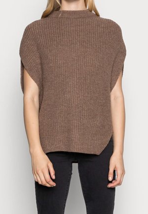 Kvinde iført en løs, ribbet, brun kortærmet sweater med høj hals og sorte tætsiddende bukser.