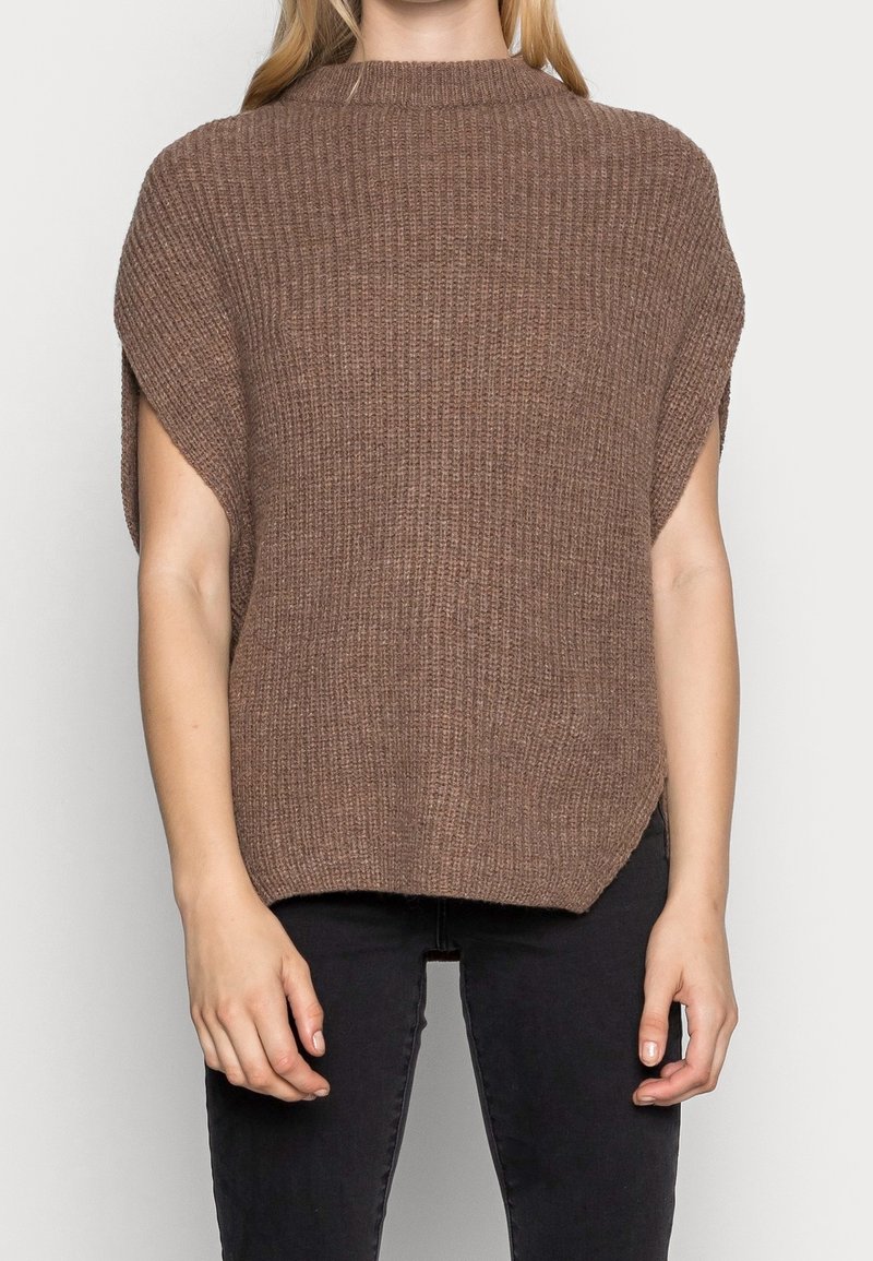 Femme portant un pull marron à manches courtes, ample et côtelé, avec un col montant et un pantalon noir ajusté.