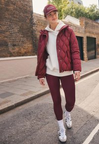 Bordeauxrote gesteppte Jacke, weißer Hoodie, weinrote Leggings und weiße Sneaker mit grauen Akzenten. Die Kappe hat eingestickten Text. Lässiger Streetwear-Stil.