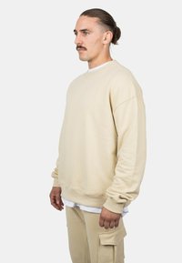 Felpa beige con vestibilità rilassata, maniche lunghe e polsini a costine. Indossata sopra una camicia bianca, abbinata a pantaloni cargo beige.