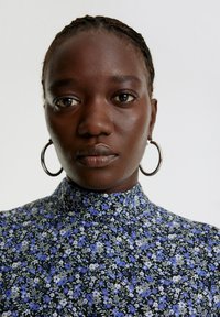 Blumenbluse mit hohem Kragen in Schwarz, verziert mit blauen und weißen Blumen. Das Model trägt große silberne Creolen und hat geflochtenes Haar.