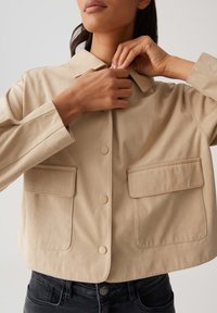 Giacca beige cropped realizzata in morbido tessuto, con colletto, due tasche frontali e chiusure a bottoni. Abbinata a jeans neri.