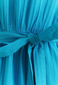 Turkoosi mekko, joka on valmistettu kevyestä kankaasta. Mekossa on kokoontunut vyötärö, jonka ympärillä on solmittava vyö, teksturoidut taitteet ja sileä viimeistely.