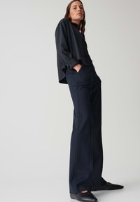 Zwarte, lange mouwen shirt onder een donkere blazer, gecombineerd met hooggewaiste, wijde zwarte broek. Glad materiaal met een getailleerd ontwerp.