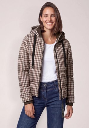 Mujer sonriente lleva chaqueta acolchada de cuadros marrón y beige con capucha, camisa blanca y pantalones vaqueros azules sobre un fondo liso.