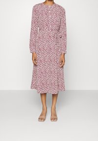 Donna che indossa un vestito a maniche lunghe, lungo fino al ginocchio, con un motivo astratto rosso e bianco e sandali beige con tacco a blocco aperti, in piedi su uno sfondo semplice.