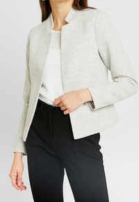 Femme portant un blazer gris clair texturé sur une blouse blanche avec un pantalon noir sur mesure, sur un fond uni.