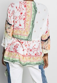 Žena v volném bílém topu s multibarevnými květinovými a paisley vzory, doplněná bílými kalhotami s modrými denimovými bočními panely.