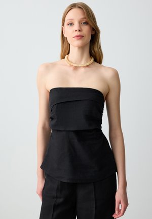 Donna con capelli castano chiaro lisci, indossa un top nero senza spalline, pantaloni neri abbinati e una collana choker dorata, in piedi contro uno sfondo grigio uniforme.