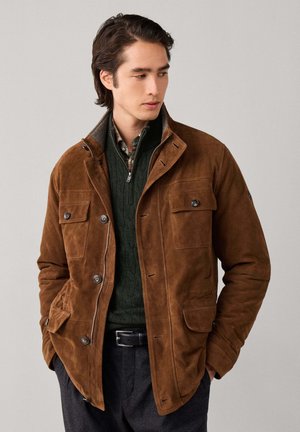 Young man looking sideways wearing a brown suede jacket, green cable-knit sweater, and checked shirt with hands in pockets.