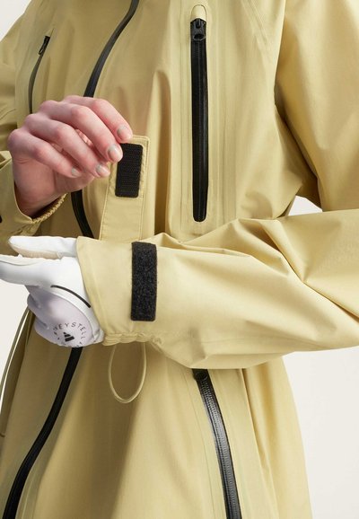 Chaqueta impermeable de color amarillo claro con bolsillos con cremallera negra y acentos de Velcro. Presenta puños elásticos y un cordón ajustable en la cintura.