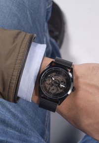 Orologio con cinturino in rete nera e quadrante rotondo, che presenta un design trasparente e più quadranti secondari, indossato su un polso con una giacca beige.