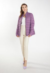 Lilla dunjakke med horisontalt quiltet design, lynlås lukning og opretstående krave. Kombineret med beige tætsiddende bukser og lavendel hæle.