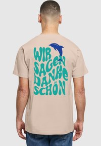 Beige t-paita, jossa on rohkea turkoosi ja sininen teksti "WIR SAGEN DANKE SCHÖN" delfiinin grafiikalla. Lyhyet hihat ja rento malli.