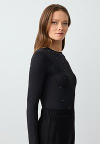 Top nero a maniche lunghe con una silhouette aderente e decorazioni di strass distribuite sulla parte anteriore. Il tessuto sembra liscio ed elasticizzato.