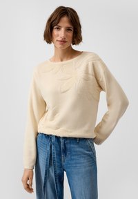 Frau mit kurzem braunem Haar, die einen cremefarbenen, floral strukturierten Pullover und hoch taillierte blaue Jeans mit einem gebundenen Jeansgürtel trägt, steht vor einem einfarbigen Hintergrund.