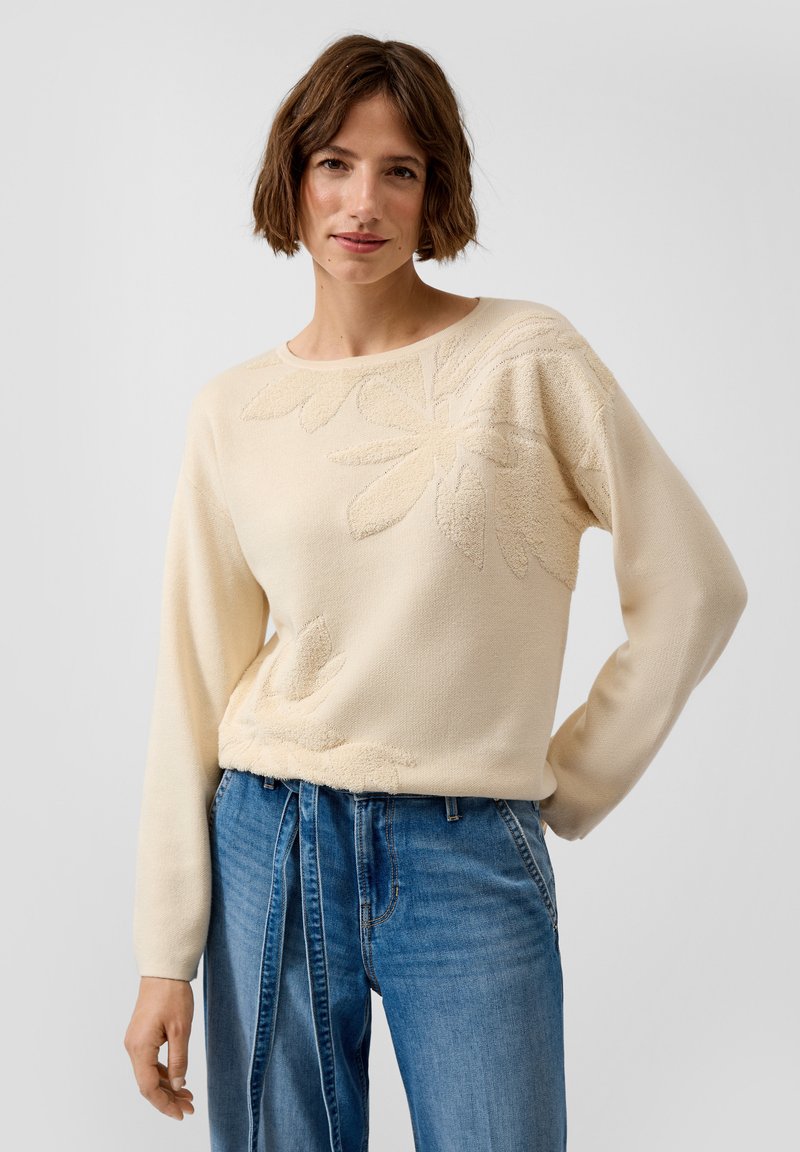 Frau mit kurzem braunem Haar, die einen cremefarbenen, floral strukturierten Pullover und hoch taillierte blaue Jeans mit einem gebundenen Jeansgürtel trägt, steht vor einem einfarbigen Hintergrund.