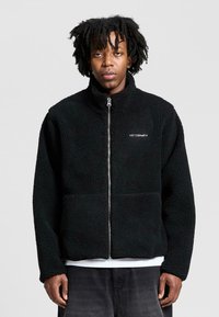 Mustat fleece-take, joissa on vetoketju edessä, korkea kaulus ja teksturoitu kangas. Vasemmassa rinnassa on 'RETURNS'-logo. Standardi mitoitus.