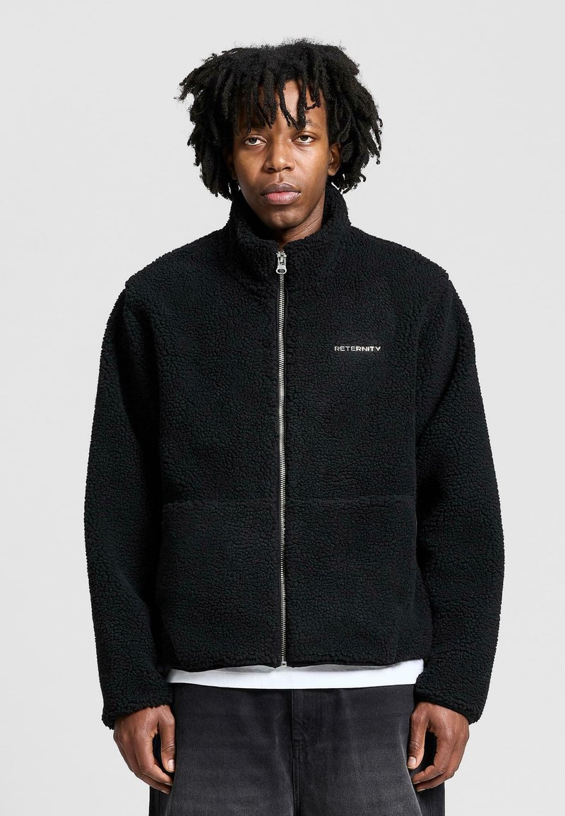 Mustat fleece-take, joissa on vetoketju edessä, korkea kaulus ja teksturoitu kangas. Vasemmassa rinnassa on 'RETURNS'-logo. Standardi mitoitus.