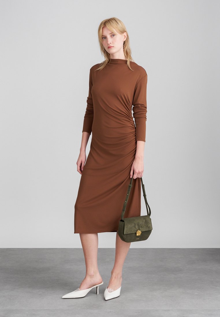Robe longue à manches longues, marron, avec fronces sur les côtés, arrivant au genou. Portée avec des mules blanches et un sac à main vert olive avec un accent doré. Matériau lisse.