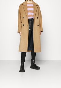 Abrigo de mezcla de lana color camel con diseño de doble botonadura, grandes botones negros, combinado con un suéter a rayas y pantalones negros. Botas gruesas negras.