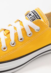 Baskets en toile jaune avec lacets blancs, œillets argentés et bordure noire. Comprend un patch logo sur la languette et une surface supérieure texturée.