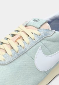 Svijetloplave traper Nike tenisice s kremastim vezicama, bijelim Swoosh logom i ružičastim detaljem na jeziku na bijeloj pozadini.