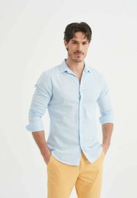 Chemise bleu clair à boutons avec des manches longues, associée à un pantalon jaune. La chemise a un col et une coupe décontractée.