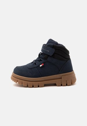Tmavomodrá členková topánka s hrubou hnedou podrážkou so vzorom, šnúrkami a suchým zipsom s logom Tommy Hilfiger.