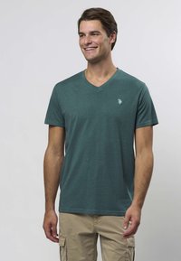 Teal V-hals t-skjorte laget av mykt stoff, med en liten logo på venstre bryst. Korte ermer og en avslappet passform.