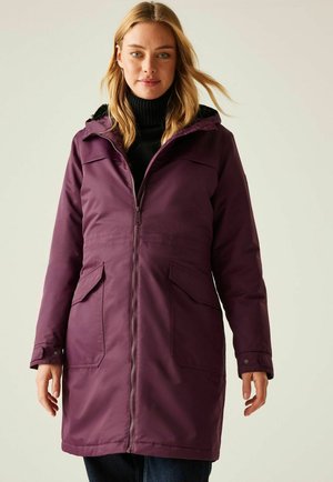 Kobieta w długim, zapinanym na zamek fioletowym zimowym płaszczu z kapturem i kieszeniami z patkami, w czarnym golfie, stojąca na jednolitym tle.