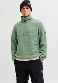 Moški oblečen v zeleni pulover z zadrgo do četrtine s logotipom "Jack & Jones", črne cargo hlače in črna kapa, stoji z rokama v žepih.