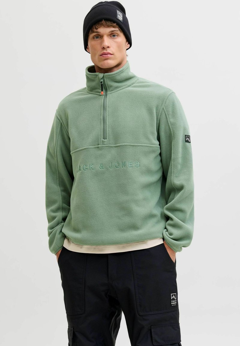 Moški oblečen v zeleni pulover z zadrgo do četrtine s logotipom "Jack & Jones", črne cargo hlače in črna kapa, stoji z rokama v žepih.