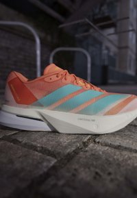 Jasnopomarańczowy but do biegania z jasnoniebieskimi paskami i białą podeszwą z napisem Lightstrike Pro na powierzchni z kostki brukowej w miejskim otoczeniu.