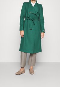 Lange, groene wollen jas met een wikkelontwerp, een strik taille en een grote kraag; heeft zilveren hardwareaccenten en een soepele textuur.