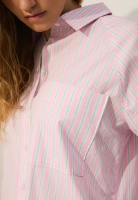 Lyserød skjorte med knapper, der har lodrette striber i lyseblå og grøn, med en brystlomme og en blød, glat tekstur.