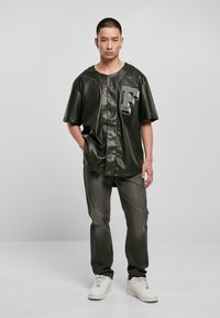 Sort læder t-shirt med korte ærmer og perforeret tekstur, udsmykket med et stort metallisk bogstav "F" på brystet, kombineret med grå jeans og hvide sneakers.