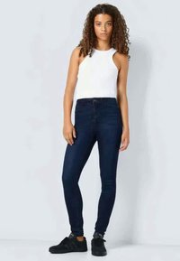 Σκούρο μπλε στενά τζιν με ψηλή μέση, συνδυασμένα με ένα εφαρμοστό λευκό αμάνικο μπλουζάκι. Το μοντέλο φοράει μαύρα αθλητικά παπούτσια. Λείος υφασμάτινος σπασμένος καμβάς.