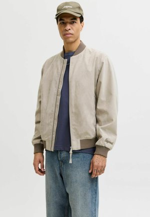 Mand iført beige bomberjakke, navyblå skjorte, lyseblå jeans og olivengrøn kasket, stående foran en ensfarvet lys baggrund.