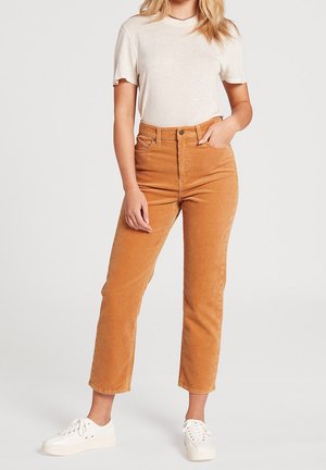 Kobieta w beżowej bluzce z krótkim rękawem, wysokich brązowych spodniach sztruksowych i białych trampkach, stoi z jedną ręką w kieszeni.