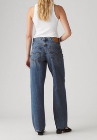 Femme aux cheveux blonds portant un jean bleu taille haute droit et un haut blanc sans manches, debout avec une main sur la hanche.
