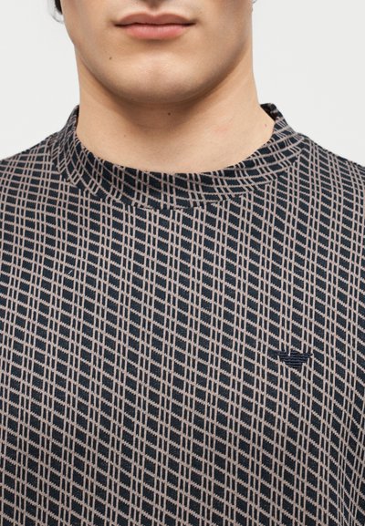 Κοντινό πλάνο ενός ατόμου που φοράει πουκάμισο με καρό σχέδιο και ψηλό γιακά, με ένα μικρό σκούρο κεντημένο λογότυπο στο στήθος.