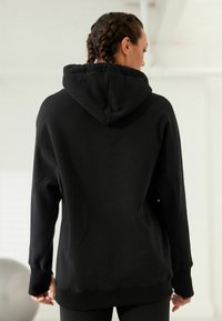 Musta huppari, jossa on suuri huppu, ribbikudottu hihansuu ja helma. Silkkinen pinta, löysä istuvuus ja minimalistinen muotoilu. Näkyy takaa.