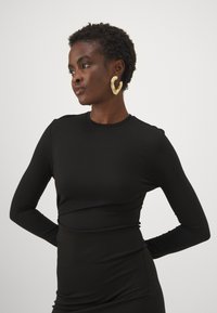 Melnā garo piedurkņu blūze, kas izgatavota no elastīga auduma ar pieguļošu dizainu, ar apaļu dekoltē un gludu tekstūru. Aksesuārs - zelta cilpas auskari.