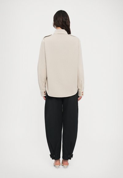 Μπεζ πουκάμισο με κουμπιά και άνετη εφαρμογή, συνδυασμένο με φαρδιά μαύρη παντελόνα με ρυθμιζόμενες μανσέτες και διακριτικές μεταλλικές λεπτομέρειες.