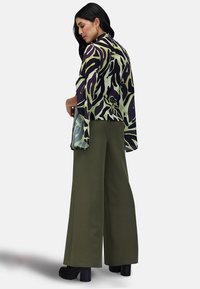 Blusa trasparente e patterned nei toni del verde e del viola, abbinata a pantaloni larghi olive a tinta unita e scarpe con zeppa nere. Maniche lunghe, design fluido.
