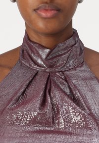 Plum metalizuota halter tipo palaidinė su klostuotu dizainu. Lygi tekstūra ir blizgus paviršius, pasižymintis siauromis vertikaliomis juostelėmis. Artimas vaizdas, akcentuojantis kaklą.